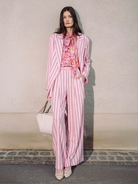 Frau im rosa Nadelstreifenanzug mit geblümter Bluse, die eine weiße Tasche in der Hand hält und helle Absätze trägt, steht an einer Wand.