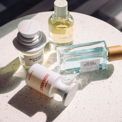 Vier verschiedene Parfümflaschen auf einer gesprenkelten weißen Oberfläche, beleuchtet von natürlichem Sonnenlicht, jede mit einzigartigen Formen und Etiketten.