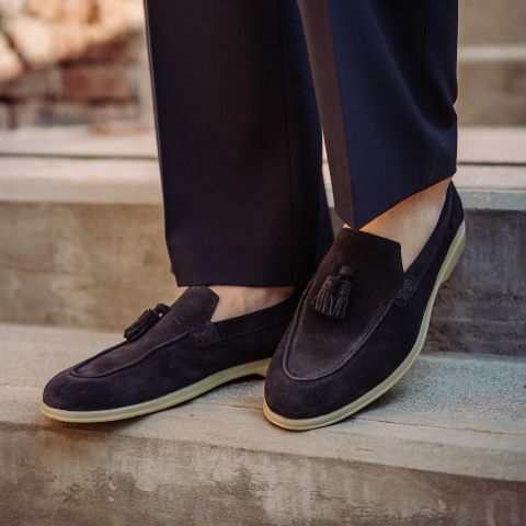 Person in dunkelnavyfarbenen Wildleder-Tassel-Slippern mit cremefarbenen Sohlen, gepaart mit marinefarbenen Hosen, stehend auf Steinstufen.