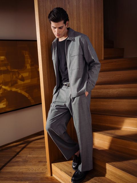 Ein Mann in grauem Anzug und schwarzem Hemd steht auf einer Holztreppe, lehnt sich mit den Händen in den Taschen an das Geländer und schaut nach unten.