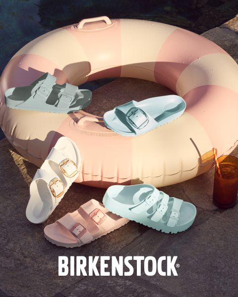 Eine Auswahl von Birkenstock-Sandalen in verschiedenen Pastellfarben wird auf einer aufblasbaren Schwimmhilfe neben einem Pool präsentiert. Ein kaltes Getränk mit Eis steht neben der Schwimmhilfe. Sonnenlicht wirft Schatten auf die Betonoberfläche.