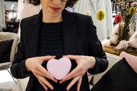 Eine Frau mit dunklen Haaren und Brille hält eine kleine, herzförmige rosa Handtasche vor ihrer Brust. Sie trägt einen schwarzen Blazer und ein gestreiftes Hemd. Im Hintergrund hängen dekorative Elemente.