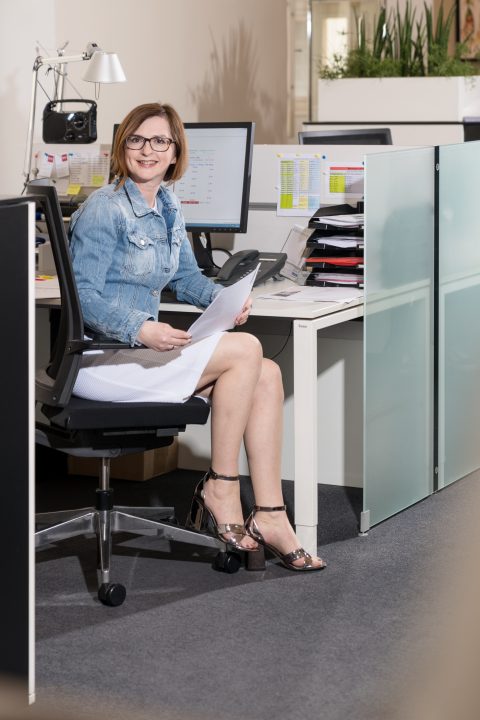 Eine Frau in Jeansjacke und weißem Kleid sitzt an einem Schreibtisch, hält Papiere in der Hand und lächelt. Der Schreibtisch ist mit einem Monitor, einer Tastatur und geordneten Aktenstapeln ausgestattet. Sie trägt eine Brille und glänzende Sandalen mit hohen Absätzen.