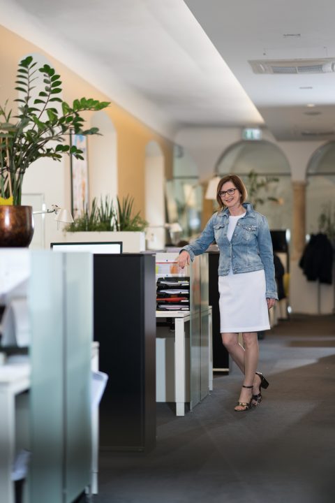 Eine Frau in weißem Kleid und Jeansjacke steht in einem Büroflur und lehnt an einem Aktenschrank mit offenen Schubladen. Der Raum ist modern, mit Topfpflanzen und gewölbten Türen im Hintergrund.