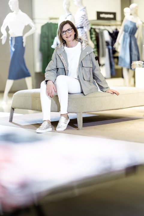 Eine Frau in grauer Jacke und weißer Hose sitzt auf einer Bank in einem Bekleidungsgeschäft. Im Hintergrund sind Schaufensterpuppen zu sehen, die verschiedene Outfits präsentieren.