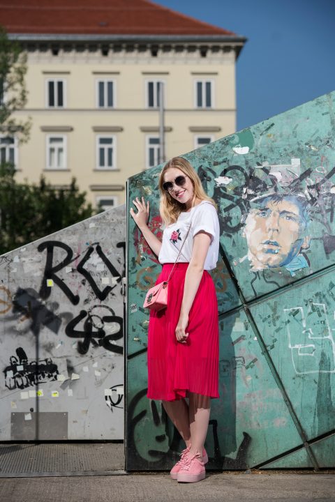 Eine Frau mit Sonnenbrille und weißem T-Shirt steht lächelnd vor einer mit Graffiti bedeckten Wand. Sie trägt einen roten Rock und rosa Schuhe, dazu eine rosa Handtasche. Im Hintergrund sind ein Gebäude und ein klarer blauer Himmel zu sehen.