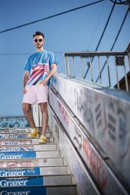 Eine Person mit Sonnenbrille und buntem Hemd steht selbstbewusst auf einer mit Graffiti besprühten Treppe unter einem klaren blauen Himmel. Ihr lässiger Look umfasst hellrosa Shorts und gelbe Schuhe und verleiht der städtischen Umgebung Lebendigkeit.