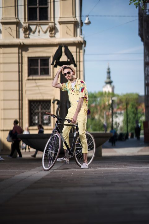 Ein Mann in gelbem Outfit und Hut posiert mit einem Fahrrad auf einer gepflasterten Straße. Er steht vor einem historischen Gebäude, im Hintergrund sind Menschen und Bäume zu sehen. Es ist ein sonniger Tag mit klarem Himmel.