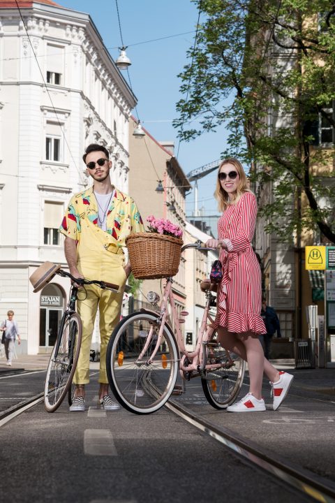 Ein Mann in einem gelben Outfit mit einem geblümten Hemd und eine Frau in einem rot gestreiften Kleid stehen mit ihren Fahrrädern auf einer Straße. Das Fahrrad der Frau hat einen Korb mit rosa Blumen. Sie sind an einem sonnigen Tag von Gebäuden und Bäumen umgeben.