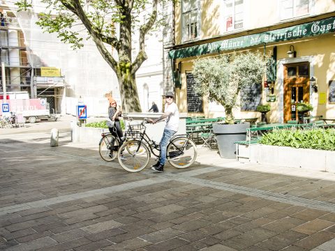 Zwei Menschen mit Fahrrädern stehen auf einer sonnenbeschienenen, von Bäumen gesäumten Straße. Sie befinden sich in der Nähe eines Gebäudes mit grünen Markisen und einem Schild auf Deutsch. Die Szene fängt eine entspannte städtische Umgebung mit Bänken, Sitzgelegenheiten im Freien und Baustellen im Hintergrund ein.