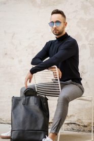 Ein Mann in schwarzem Rollkragenpullover, graukarierter Hose und Sonnenbrille sitzt auf einem weißen Stuhl vor einer hellen Wand. Neben ihm liegt eine große schwarze Ledertasche auf dem Boden.