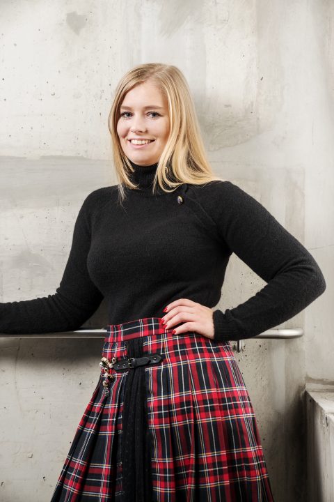 Eine Person mit langen blonden Haaren lächelt, trägt einen schwarzen Rollkragenpullover und einen rot-schwarz karierten Rock und steht vor einer Betonwand.