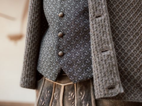 Nahaufnahme einer Person, die eine strukturierte graue Strickjacke über einer grau gemusterten Weste mit drei runden Knöpfen trägt. Der untere Teil zeigt einen Rock mit Falten und dezenten gestickten Details. Der Hintergrund ist unscharf.