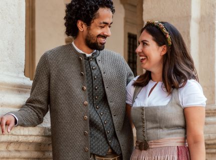 Ein Mann und eine Frau lächeln sich an. Sie sind in traditioneller bayerischer Kleidung gekleidet. Der Mann trägt eine strukturierte Jacke und ein Hemd und die Frau ein Kleid mit einem geblümten Stirnband. Sie stehen neben einer hellen Säule im Freien.