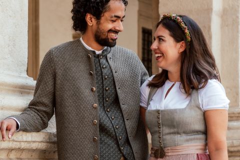 Ein Mann und eine Frau lächeln sich an. Sie sind in traditioneller bayerischer Kleidung gekleidet. Der Mann trägt eine strukturierte Jacke und ein Hemd und die Frau ein Kleid mit einem geblümten Stirnband. Sie stehen neben einer hellen Säule im Freien.