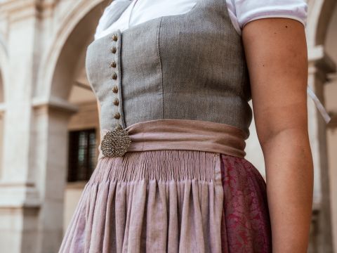 Nahaufnahme einer Person in traditioneller Kleidung mit einem taillierten grauen Mieder, verzierten Knöpfen und einem weiten Rock in Lavendel- und Rottönen. Sie stehen vor einem Hintergrund aus Steinbögen.