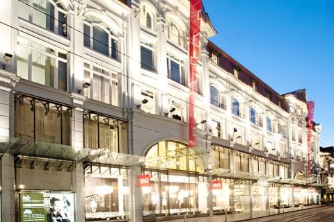 Beleuchtete Fassade eines großen, eleganten Einkaufszentrums am Abend, mit gewölbten Fenstern und Glasdächern. Die Straße ist leer, und die Schaufenster sind hell beleuchtet.