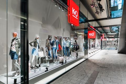 Ein Schaufenster eines Modegeschäfts, in dem Schaufensterpuppen in trendigen Freizeitoutfits, darunter Jacken und Jeansshorts, ausgestellt sind. Das Geschäft hat große Glasfenster und befindet sich in einem modernen Einkaufszentrum mit eleganter Architektur.