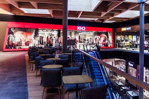 Das Interieur eines modernen Einkaufszentrums mit einem rot beleuchteten Bekleidungsgeschäft namens K&O. Der Raum umfasst Sitzbereiche mit Stühlen und Tischen, eine Rolltreppe und verschiedene andere Schaufenster im Hintergrund.