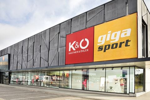 Außenansicht eines modernen Gebäudes mit großen Schildern für „Kastner & Öhler“ in Rot und „Giga Sport“ in Gelb. Im Erdgeschoss sind große Schaufenster mit Sport- und Modeartikeln zu sehen. Der Himmel ist bedeckt.