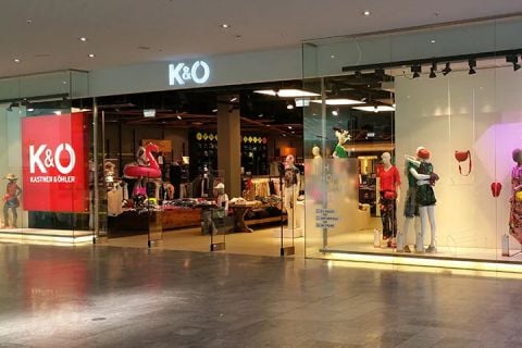 Schaufenster von Kastner & Öhler mit großen Glasfenstern, in denen bekleidete Schaufensterpuppen zu sehen sind. Im Ladeninneren sind Kleiderständer und eine Auswahl an Bekleidung zu sehen. Das K&O-Logo leuchtet deutlich sichtbar über dem Eingang.