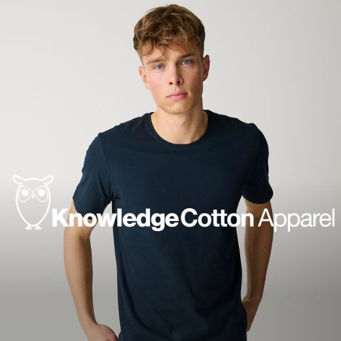 Ein junger Mann in einem schlichten dunklen T-Shirt steht vor einem hellen Hintergrund, auf dem das Logo und der Text von "Knowledge Cotton Apparel" überlagert sind.