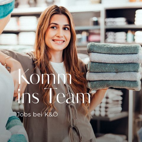 Junge Frau lächelt und hält gefaltete Handtücher in einem Geschäft mit deutschem Text darüber: "Komm ins Team. Jobs bei K&Ö." Regale mit Handtüchern im Hintergrund.