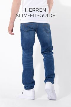 Vor einem weißen Hintergrund steht ein Mann, der blaue Slim-Fit-Jeans und weiße Sneakers trägt. Sein Oberkörper ist teilweise sichtbar und er blickt von der Kamera weg. Über ihm ist der Text „HERREN SLIM-FIT-GUIDE“ zu sehen.