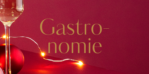 Ein Glas Weißwein und eine leuchtende Lichterkette stehen neben einem roten Weihnachtsornament vor einem leuchtend roten Hintergrund. Das Wort „Gastronomie“ ist in goldener Schrift geschrieben.