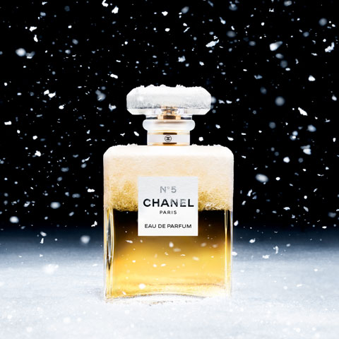 Abgebildet ist eine Flasche Chanel No. 5 Parfüm, deren Deckel und Oberseite mit Schnee bedeckt sind. Um sie herum fallen Schneeflocken herab, die vor einem schwarzen Hintergrund eine winterliche Atmosphäre erzeugen.