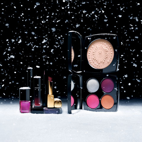 Eine Präsentation von Make-up-Produkten auf einer schneebedeckten Oberfläche, darunter Nagellack, Lippenstift und ein Kompaktset mit vier bunten Farbtönen. Schneeflocken fallen vor einem dunklen Hintergrund.