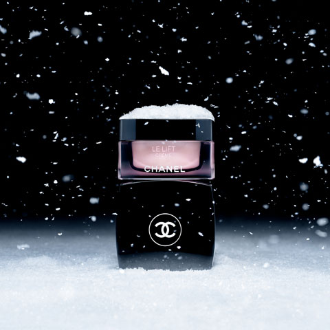 Ein Chanel Le Lift Cremebehälter, umgeben von fallenden Schneeflocken, auf denen Schnee liegt. Der Hintergrund ist dunkel, was den Kontrast zum weißen Schnee und dem glänzenden Schwarz und Pink der Verpackung verstärkt.