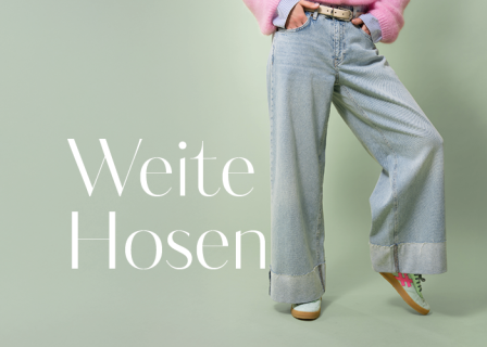 Person in Jeans mit weitem Bein und rosa Pullover posiert vor hellgrünem Hintergrund. Links ist der Schriftzug „Weite Hosen“ zu sehen. Grüne und rosa Turnschuhe sind zu sehen.