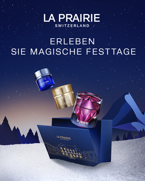 Ein Werbebild für La Prairie zeigt drei luxuriöse Hautpflegeprodukte – zwei Tiegel und eine Flasche – schwebend über einer dunkelblauen Geschenkbox, vor einer Kulisse aus schneebedeckten Bergen und einem sternenklaren Nachthimmel. Der Text lautet: „Erleben Sie magische Festtage.“