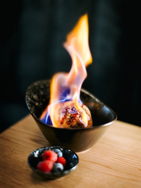 Ein Gericht mit einer Kugel Dessert, das in einer schwarzen Schüssel in leuchtenden Flammen brennt und auf einer Holzoberfläche steht. Dazu gibt es eine kleine Schale mit gemischten Beeren, darunter Himbeeren und Blaubeeren.