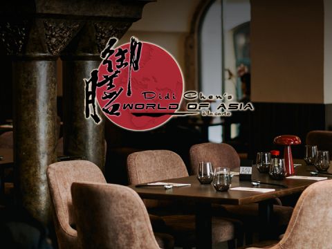 Ein schwach beleuchtetes Restaurant-Interieur mit braunen Stühlen und gedeckten Tischen mit Weingläsern und einer kleinen Lampe. Im Hintergrund ist ein Schild in Rot und Schwarz mit der Aufschrift „Didi Chens Welt Asiens à la carte“ mit stilisierten asiatischen Schriftzeichen zu sehen.