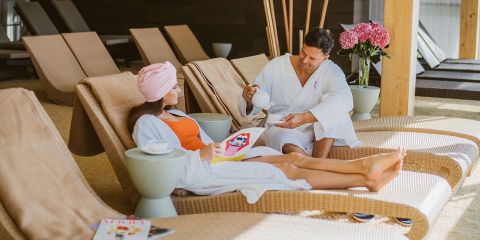 Ein Mann und eine Frau in weißen Bademänteln entspannen auf Liegestühlen in einem Spa. Der Mann schenkt Tee ein und die Frau hält eine Zeitschrift in der Hand. Im Hintergrund sind ein rosa Blumenarrangement und weitere Liegestühle zu sehen.