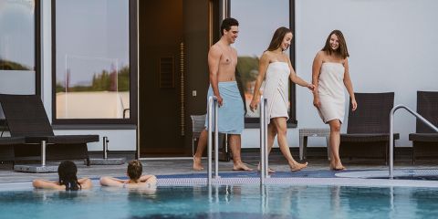 Drei in Handtücher gehüllte Personen laufen lächelnd und unterhaltend an einem Pool entlang, während zwei andere im Wasser entspannen. Mit Liegestühlen und einem modernen Gebäude im Hintergrund wirkt die Umgebung ruhig.