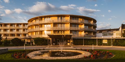 Ein modernes, rundes Hotelgebäude mit mehreren Balkonen, getaucht in das warme Licht des Sonnenuntergangs. Im Vordergrund ist ein runder Brunnen zu sehen, umgeben von blühenden Blumen und einem gepflegten Garten. Der Himmel ist teilweise bewölkt.