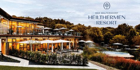 Außenansicht des Heiltherme Bad Waltersdorf Resorts mit modernem Design, großen Fenstern und Sitzbereichen im Freien mit Sonnenschirmen. Umgeben von üppigem Grün ist die Szene vor einer Kulisse aus Bäumen unter einem teilweise bewölkten Himmel angesiedelt.