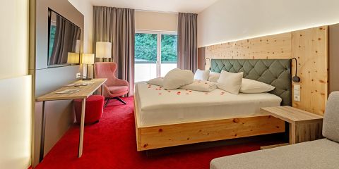 Ein gemütliches Hotelzimmer mit einem Doppelbett aus Holz, das mit Rosenblättern geschmückt ist. An der Wand hängt ein großer Fernseher, links steht ein Schreibtisch mit Stuhl. Natürliches Licht strömt durch ein Fenster mit grauen Vorhängen herein. Teppich und Stuhl sind rot.