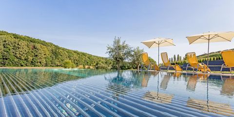 Eine ruhige Poolszene mit einem Infinity-Pool, in dem sich der klare blaue Himmel spiegelt. Gelbe Liegestühle und Sonnenschirme säumen den Poolrand neben üppigen grünen Bäumen. Das Wasser ist ruhig und bietet eine friedliche und entspannende Atmosphäre.