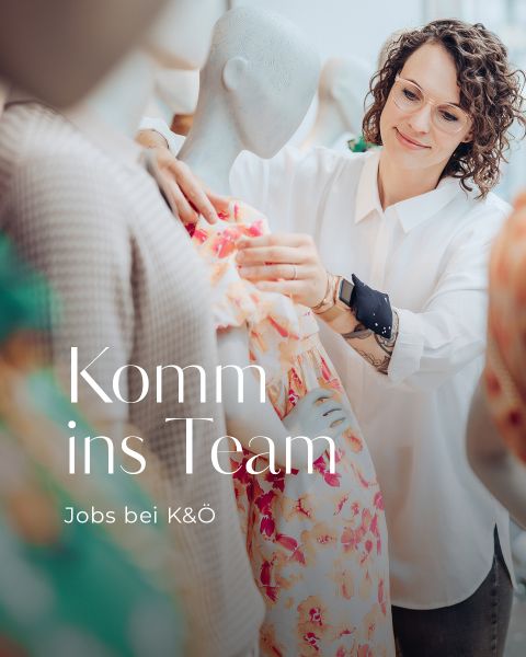 Eine Frau im weißen Hemd lächelt, während sie an einer Schaufensterpuppe Kleidung zurechtrückt. Auf dem Bild ist der Text zu sehen: „Komm ins Team“ und „Jobs bei K&O“.