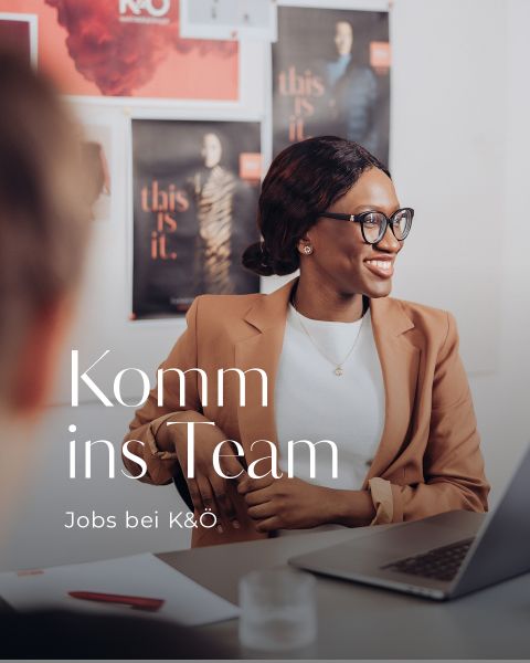 Eine Person mit Brille und braunem Blazer sitzt lächelnd an einem Tisch mit einem Laptop. Im Hintergrund hängen Plakate an der Wand. Der Text auf dem Bild lautet „Komm ins Team“ und „Jobs bei K&O“.