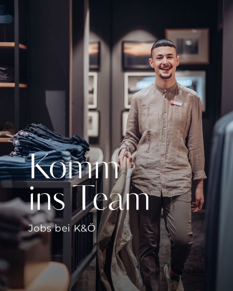 Eine lächelnde Person in legerer Kleidung geht durch ein Bekleidungsgeschäft. Im Hintergrund sind gefaltete Jeans zu sehen. Auf dem Bild steht: „Komm ins Team. Jobs bei K&O.“