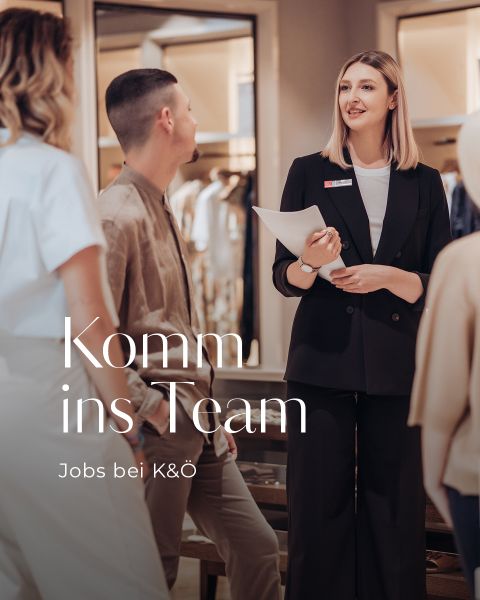 Eine Frau im schwarzen Anzug spricht mit zwei Personen in einem Büro. Sie hält Papiere in der Hand. Der Text lautet „Komm ins Team“ und „Jobs bei K&Ö“.