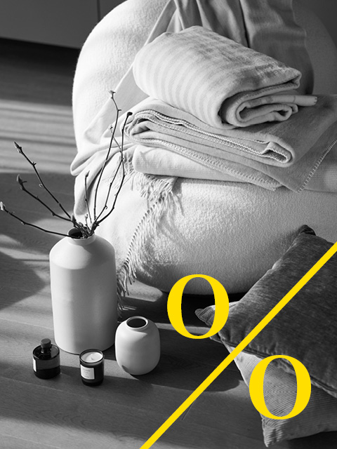 Eine gemütliche Szene mit gefalteten Decken, einem Sitzsack, Vasen mit Zweigen und Kerzen auf einem Holzboden bei natürlichem Licht.