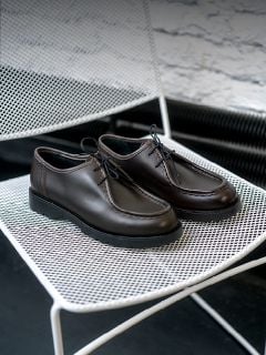 Person in hochgekrempelten Jeans steht auf sandigem Boden in der Nähe von Kakteen und trägt weiße Turnschuhe mit beigen und braunen Akzenten und einem "V"-Logo an der Seite.