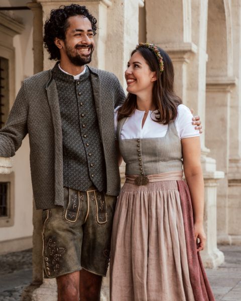 Ein Mann und eine Frau lächeln sich an und tragen traditionelle bayerische Kleidung. Der Mann hat einen Bart, lockiges Haar und trägt eine Weste und bestickte Lederhosen. Die Frau hat ein Blumenstirnband und ein Kleid mit Weste und weitem Rock. Sie stehen vor einem Gebäude.