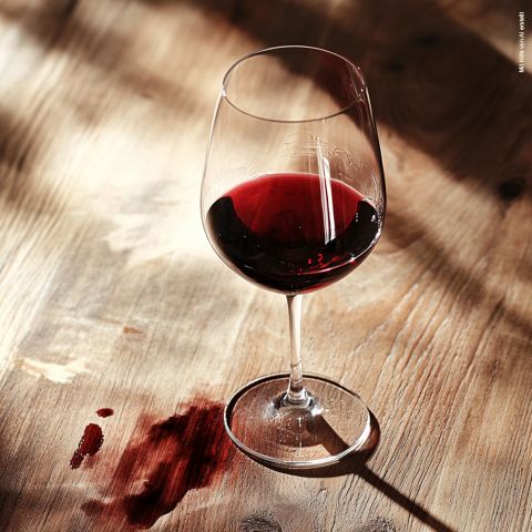 Ein Glas Rotwein steht auf einem Holztisch und wirft einen Schatten. In der Nähe sorgen ein paar Weinflecken für einen roten Farbtupfer auf der Holzoberfläche, der durch das einfallende Sonnenlicht noch hervorgehoben wird.